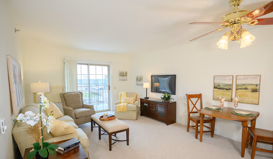 A bright and cozy living room in a senior community with a beige sofa, two armchairs, a wooden coffee table, a TV mounted on a wooden cabinet, and a small dining table with two chairs. The room has a ceiling fan with lights, wall art, a sliding glass door leading to a balcony, and soft carpeting.