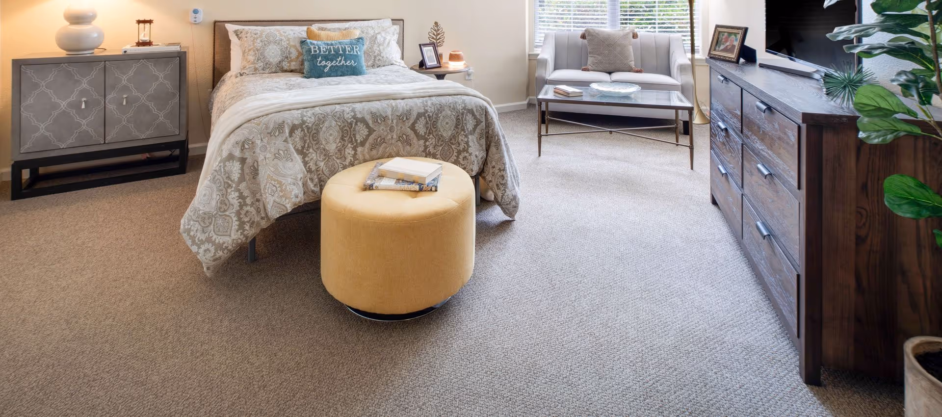 Sunlit furnished bedroom with a patterned bed, round yellow ottoman, wooden dresser, and a small seating area by the window.