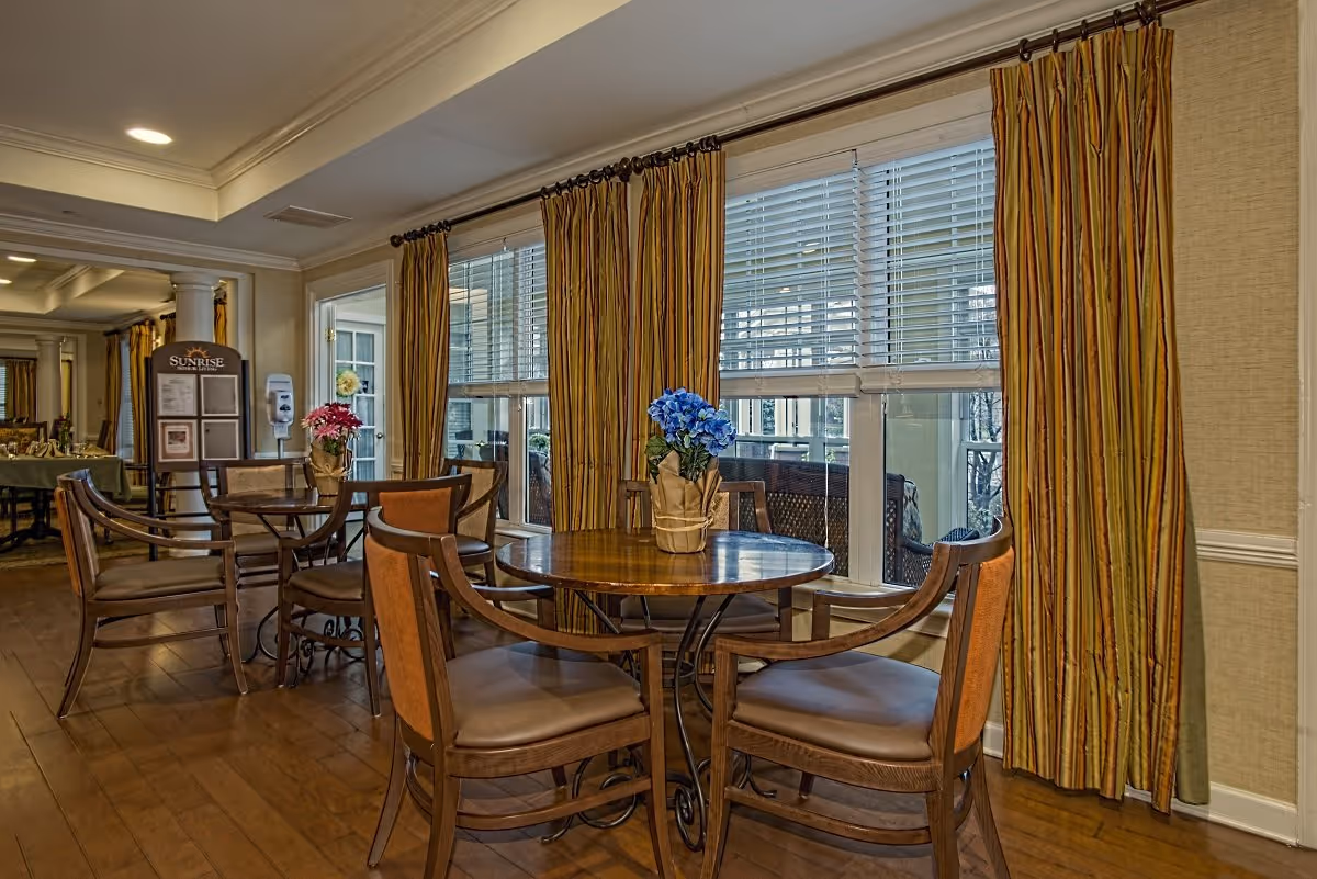 Cozy dining area with round wooden tables and chairs, flower centerpieces, and large windows with striped curtains.
