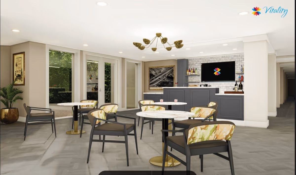 A modern common area with round tables and chairs featuring floral upholstery. There is a bar counter with bottles and a TV mounted on a white brick wall. Large windows and glass doors open to an outdoor space with greenery visible. A decorative chandelier hangs from the ceiling, and a framed picture is on the wall.