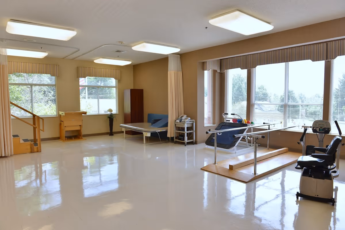 A spacious rehabilitation or physical therapy room with large windows letting in natural light. The room contains exercise equipment including parallel bars, a recumbent exercise bike, a therapy table, and a small set of stairs with a handrail. The floor is shiny and clean, and the walls are beige with large windows covered by blinds and valances.