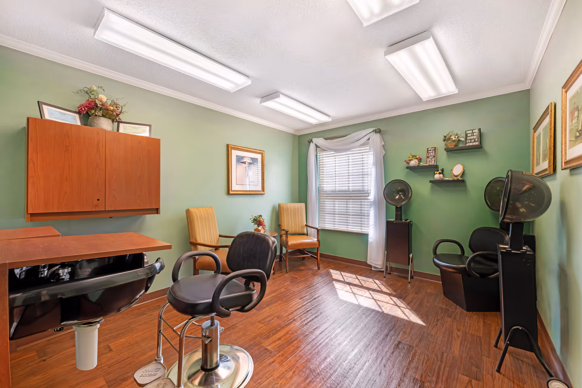 Small salon-style room with green walls, wood flooring, salon chairs and hooded hair dryers under bright ceiling lights.
