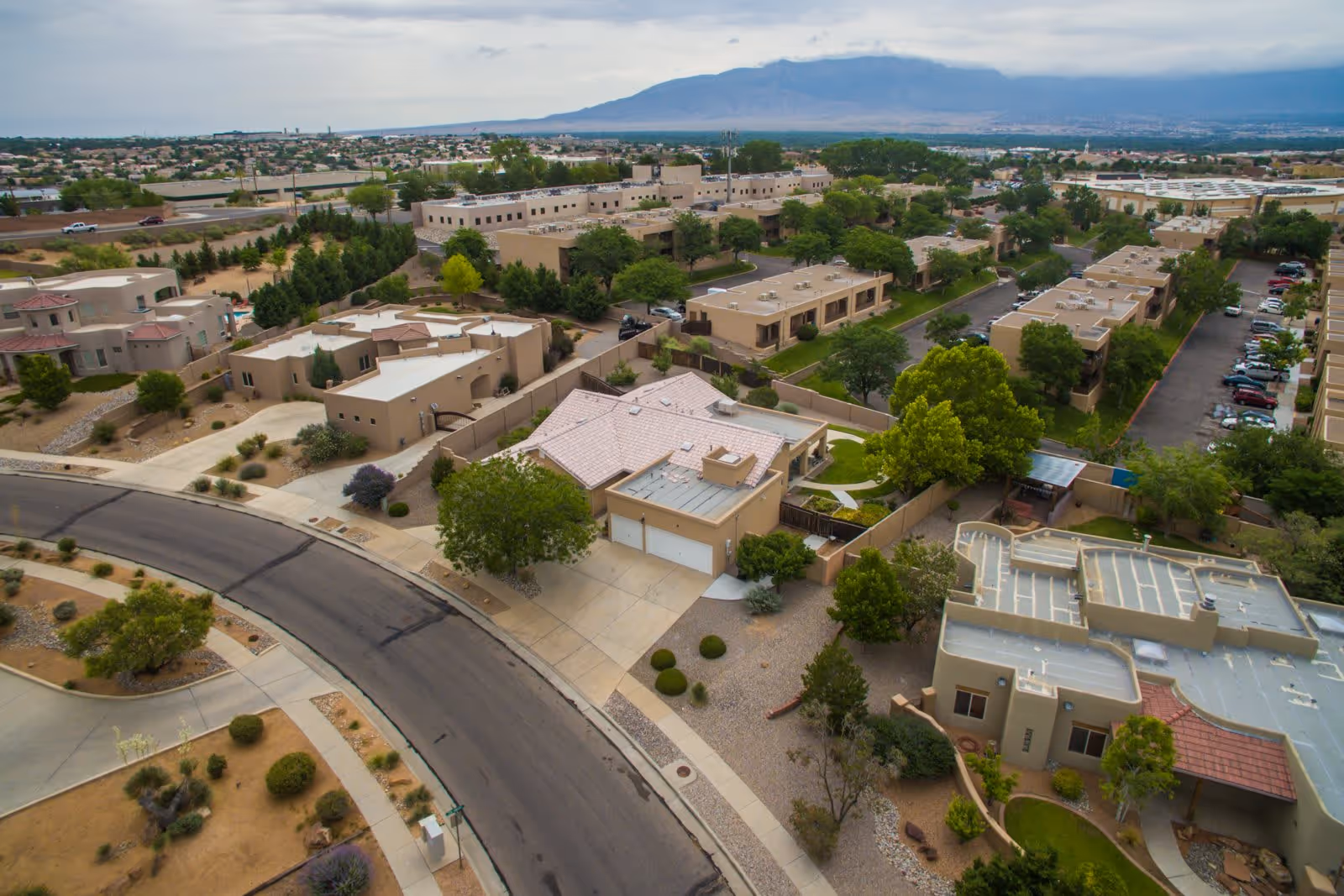 Aerial view of a residential neighborhood with multiple beige and tan buildings, trees, and parked cars. The area includes single-family homes and apartment-style buildings with a mountain range visible in the background under a cloudy sky.