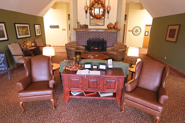 A cozy living room area with two brown leather armchairs in the foreground, a wooden console table between them, and a fireplace with autumn-themed decorations including pumpkins and a wreath above it. The room has warm lighting, patterned carpet, and green and beige walls with framed pictures and decorative items.