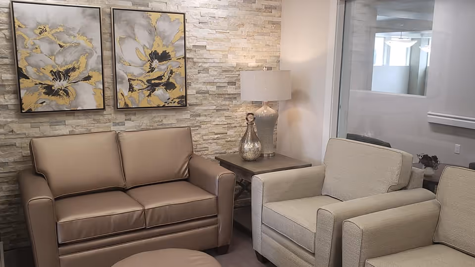Seating area with a brown leather loveseat, two beige armchairs, a side table with a lamp and decorative vases, and floral paintings on a stone accent wall.