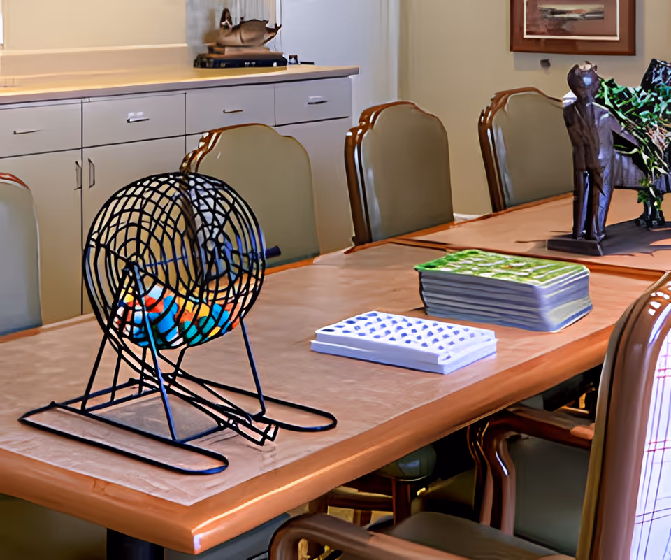 A long wooden table with padded chairs around it in a room. On the table, there is a black wire bingo cage with colorful bingo balls inside, a bingo card holder, and a stack of bingo cards. In the background, there are cabinets with drawers and a framed picture on the wall.