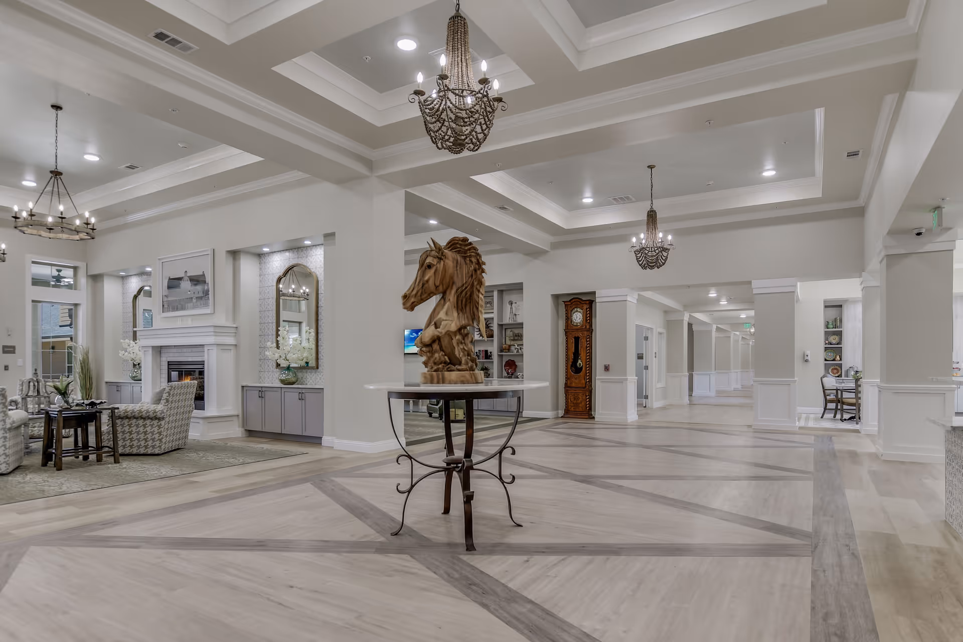 Spacious and elegant senior living facility interior with high coffered ceilings, chandeliers, a wooden horse head sculpture on a round table in the center, a seating area with armchairs and a fireplace on the left, and a long hallway leading to other rooms on the right.