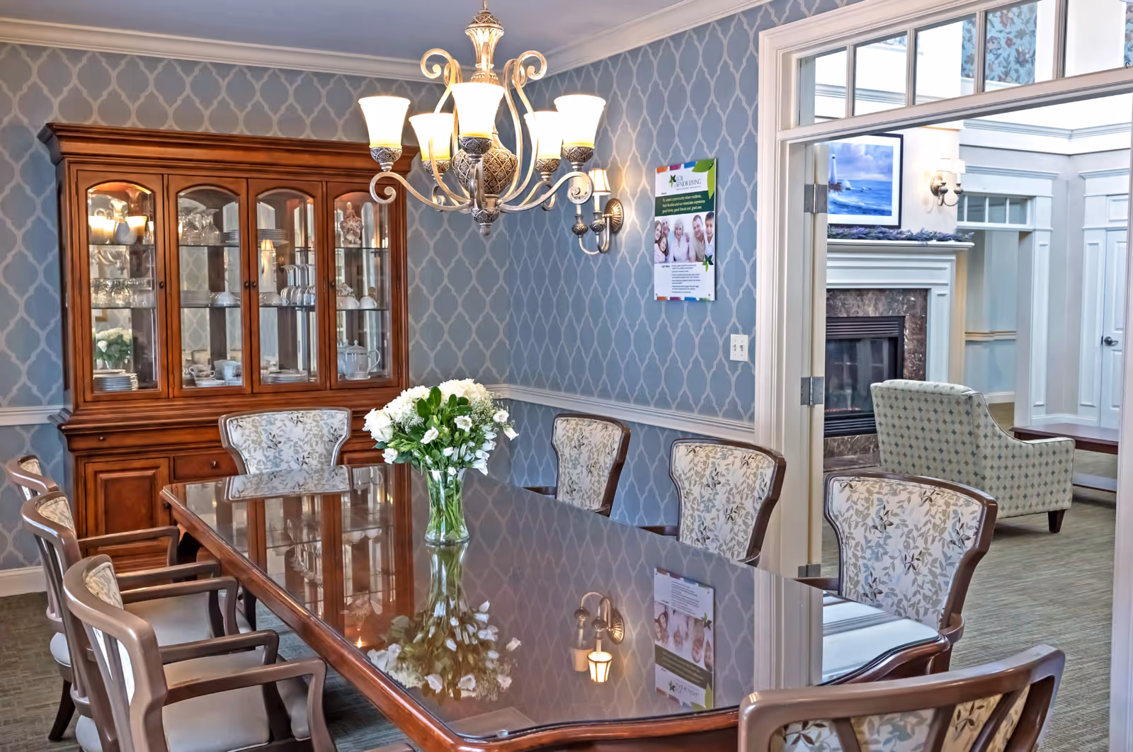 A formal dining room with a polished wooden table surrounded by eight upholstered chairs with floral patterns. A glass vase with white flowers is centered on the table. Behind the table is a wooden china cabinet displaying glassware and dishes. The walls are decorated with blue patterned wallpaper, and a chandelier with multiple lights hangs above the table. Through an open doorway, a living room area with a fireplace, armchair, and framed artwork is visible.