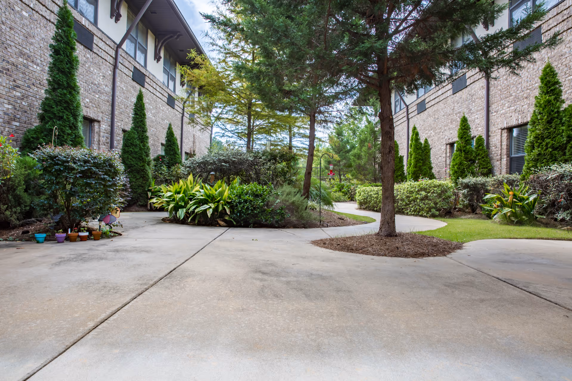 Outdoor courtyard area between two brick buildings with a concrete walkway, landscaped with various green shrubs, small trees, and potted plants. The scene is peaceful and well-maintained with a large tree in the center and a bird feeder hanging from a pole.