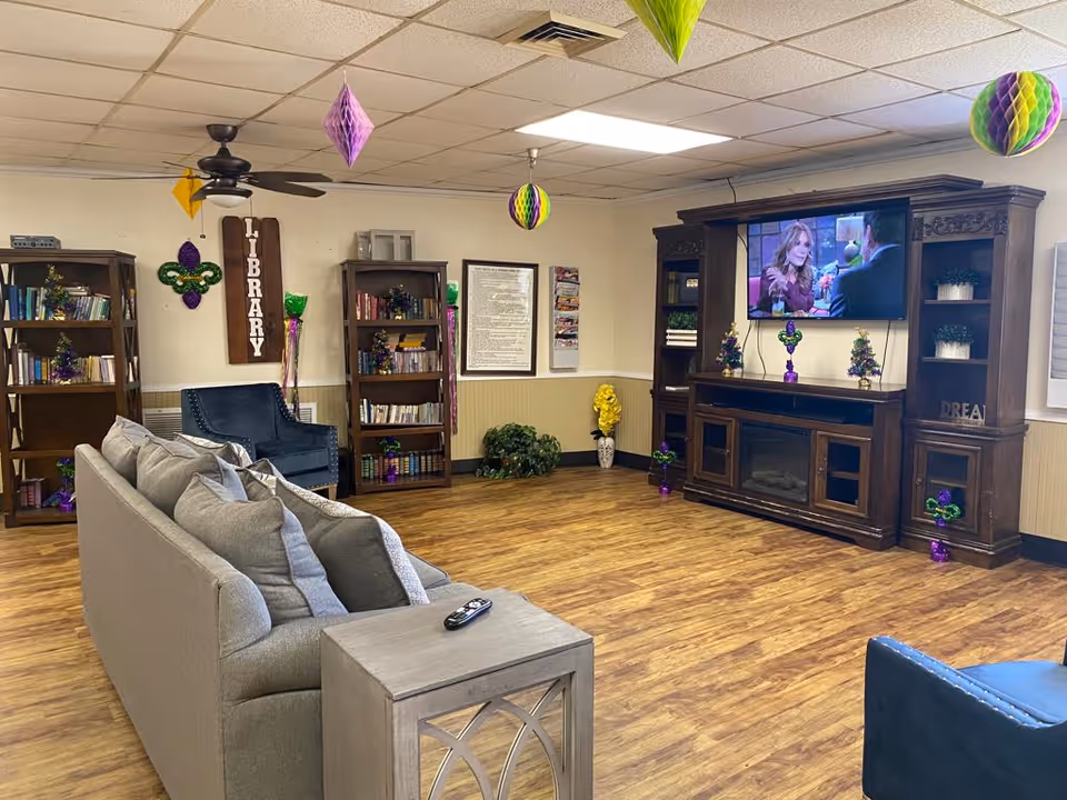 A cozy living room area with a gray couch, a blue armchair, wooden bookshelves filled with books, and a large wooden entertainment center with a flat-screen TV mounted above an electric fireplace. The room has wood flooring, ceiling tiles, a ceiling fan, and colorful hanging decorations. A wooden sign on the wall reads 'LIBRARY'.