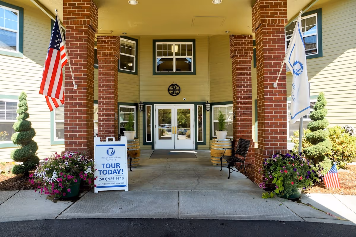 Entrance to Prestige Senior Living Riverwood featuring a covered walkway with brick pillars, two American flags, potted plants with flowers, a bench, and a sign advertising tours with contact information.