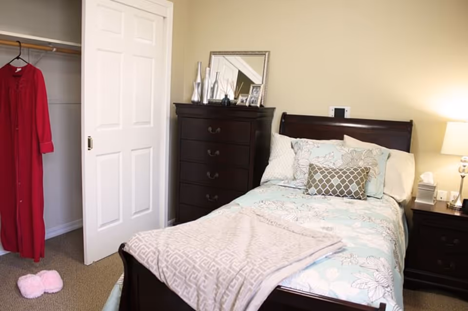 A cozy bedroom with a single bed made up with floral patterned bedding and multiple pillows. Next to the bed is a dark wooden nightstand with a lamp and tissue box. A matching dark wooden dresser with decorative items and a mirror is against the wall. An open closet door reveals a red robe hanging inside and a pair of pink slippers on the carpeted floor.
