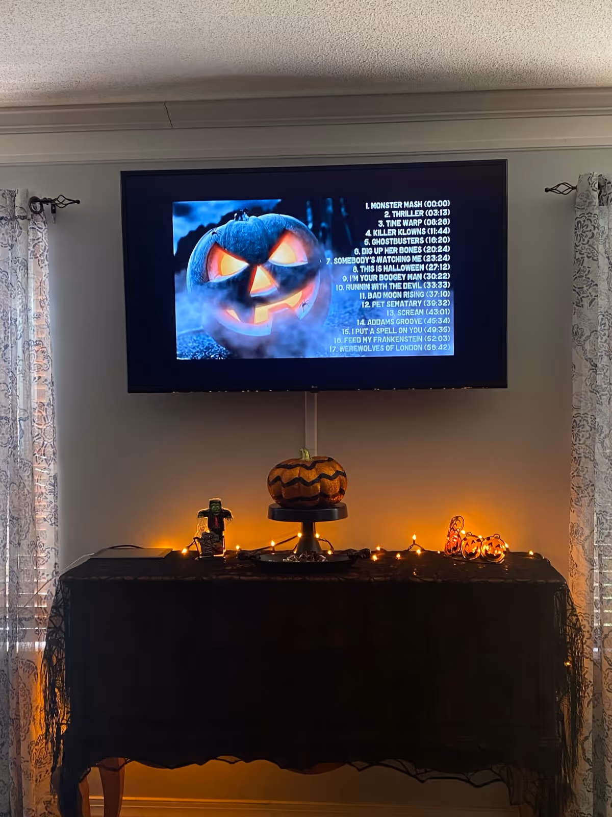 A living room area with a flat-screen TV mounted on the wall displaying a Halloween-themed playlist with a glowing jack-o'-lantern image. Below the TV is a table covered with a black cloth, decorated with a pumpkin on a stand, a small Frankenstein figure, orange string lights, and a 'Boo' sign. Light-colored curtains frame the scene on either side.