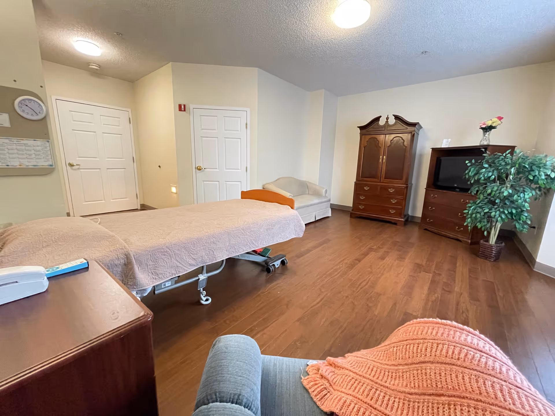 Private senior living bedroom with an adjustable bed, armchair, wooden armoire and TV on hardwood floors.