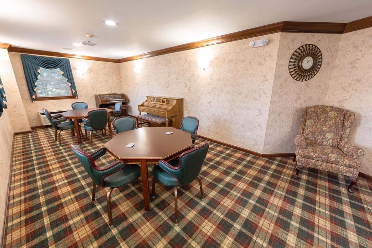 A common room with patterned carpet and beige wallpaper featuring two octagonal wooden tables surrounded by green cushioned chairs. There is a floral upholstered armchair in the corner, a wall clock, and two musical instruments including a piano and an organ against the far wall. The room has a window with green drapes and is lit by wall sconces and ceiling lights.