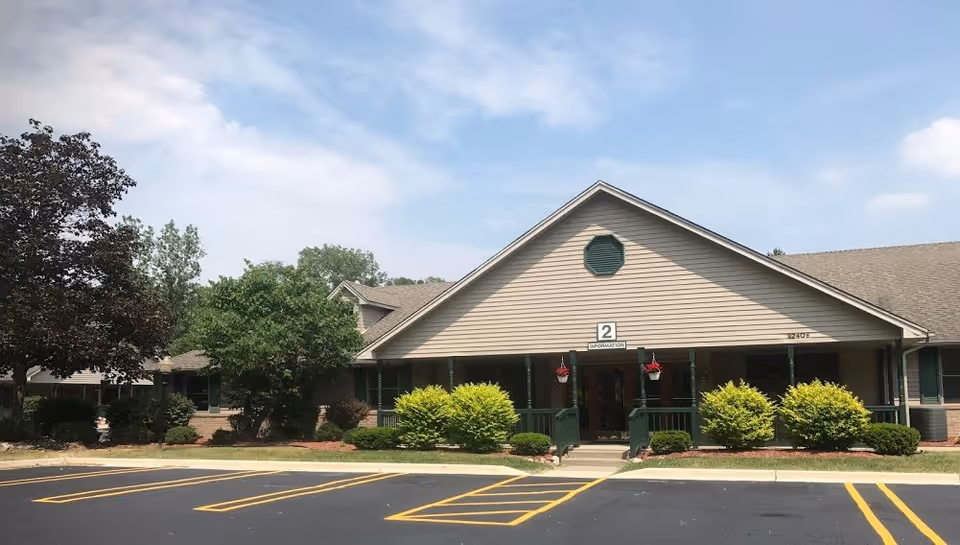 Exterior view of a single-story building with beige siding and a gable roof, surrounded by green bushes and trees under a partly cloudy sky. There is a parking lot with marked spaces in front of the building and a sign above the entrance that reads '2 INFORMATION'.
