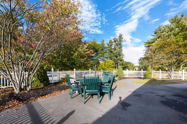 Outdoor patio with green Adirondack-style chairs around a table, a white picket fence, and trees under a blue sky.