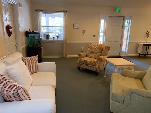A cozy living room area with a white couch adorned with striped pillows, a floral armchair, and a green armchair surrounding a small wooden coffee table. There is a window with sheer curtains, a fish tank on a stand, and a door with a heart-shaped decoration. The room has beige walls and green carpet flooring.