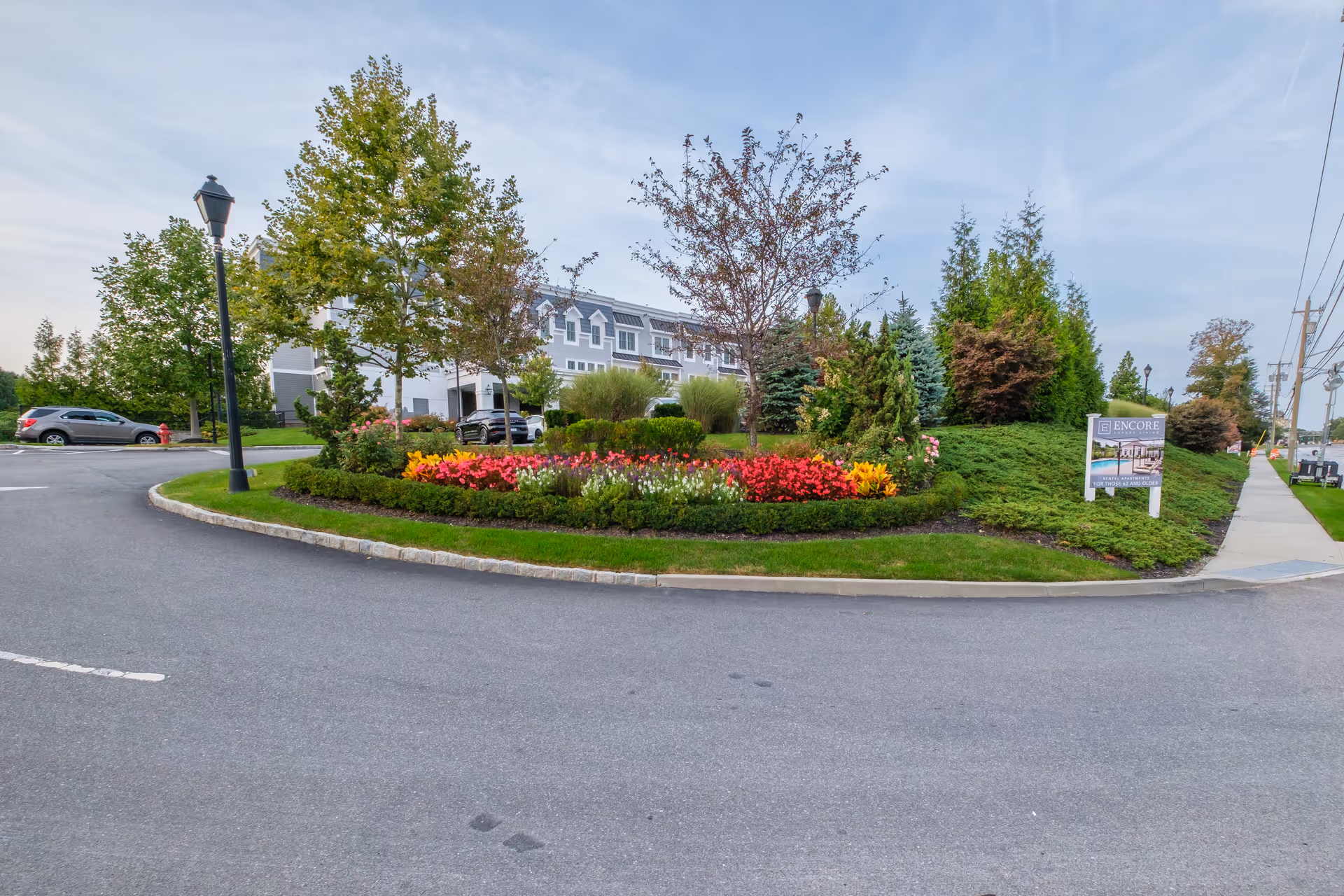 Exterior view of Encore Luxury Living facility showing a landscaped garden with colorful flowers and various trees. A paved road curves around the garden, and a sign for Encore Luxury Living is visible on the right side near the sidewalk. Several parked cars and a multi-story building are in the background under a partly cloudy sky.