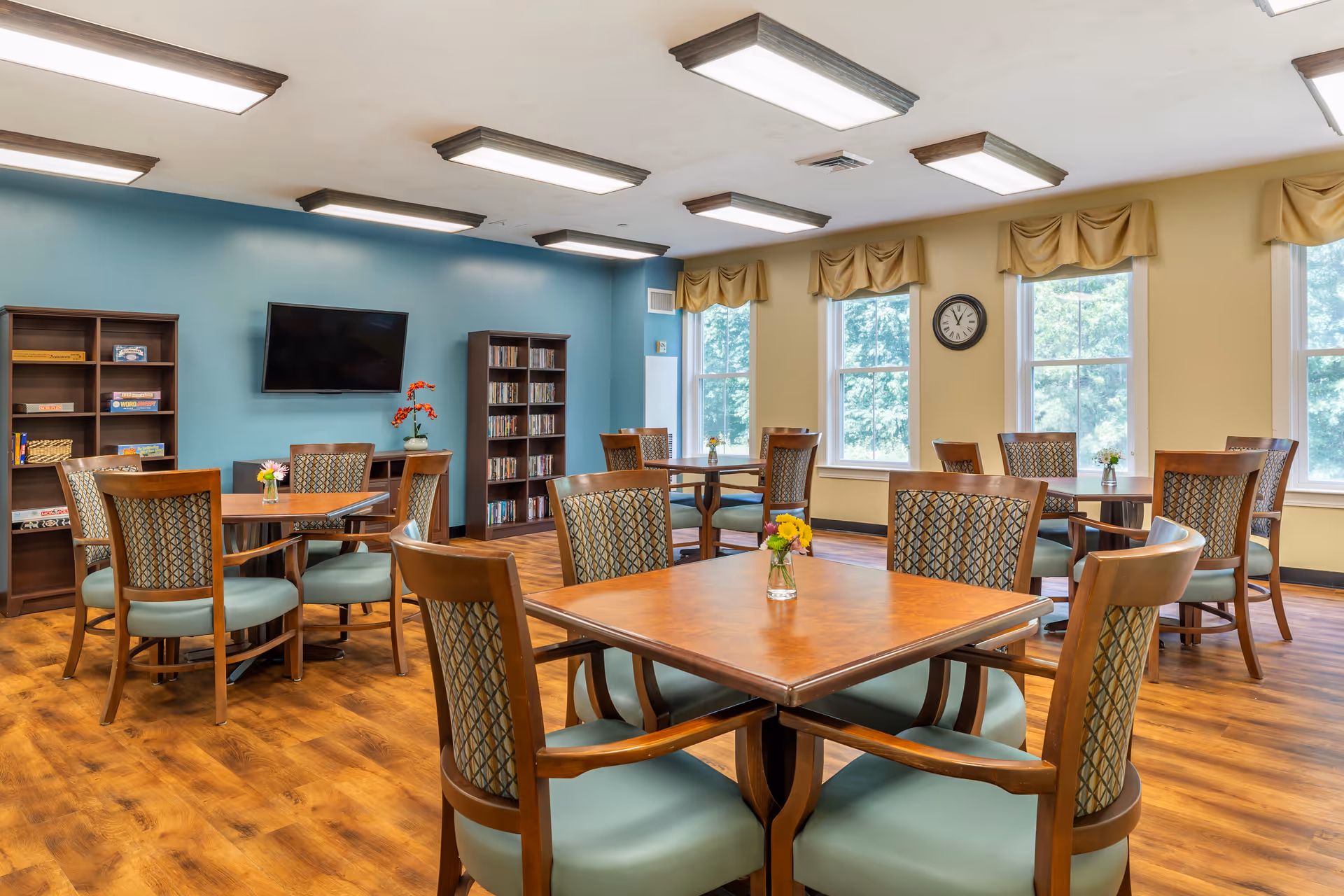 A bright and spacious common room with several wooden tables and chairs arranged neatly. The room has large windows with beige valances, allowing natural light to fill the space. There are bookshelves filled with books and board games, a wall-mounted TV, and small flower vases on each table. The walls are painted in a combination of blue and beige, and the floor has a warm wood finish.