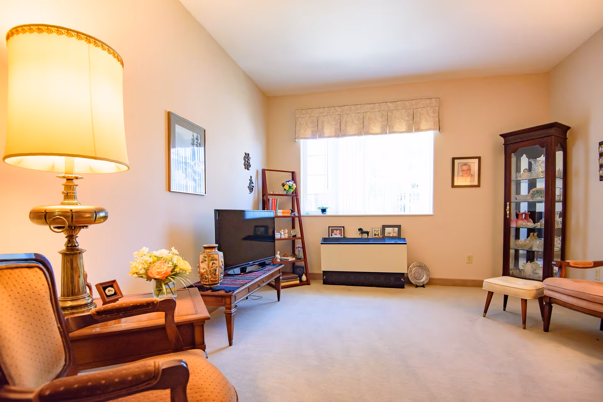 A cozy living room with beige walls and carpet, featuring a large window with a valance. The room contains a wooden side table with a brass lamp and a flower vase, two upholstered armchairs, a wooden TV stand with a flat-screen TV and decorative items, a leaning bookshelf with books and ornaments, a glass display cabinet with collectibles, and framed pictures on the walls and window ledge.