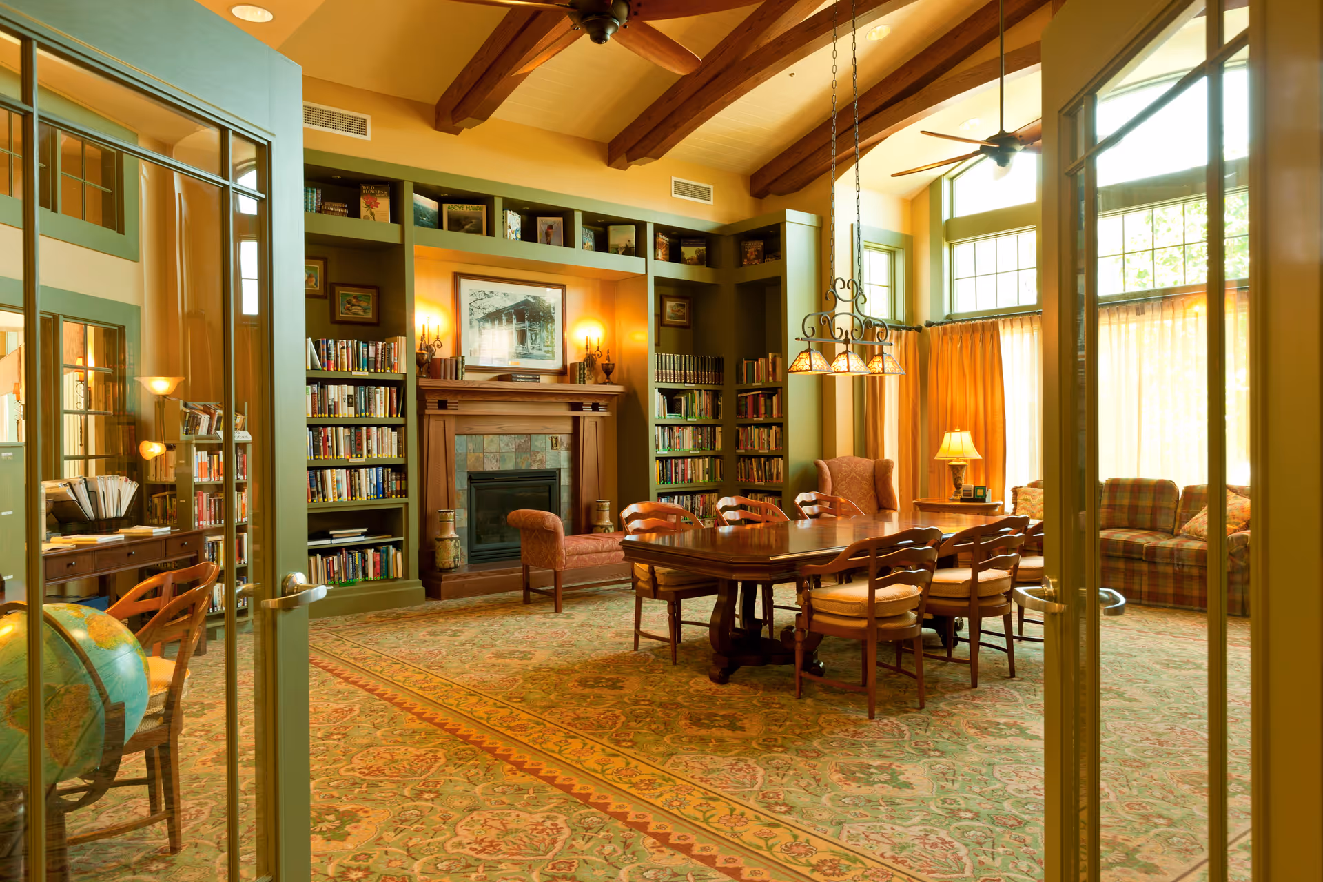 Sunlit communal living room/library with a large wooden table and chairs, built-in bookshelves, a fireplace, and comfortable seating.