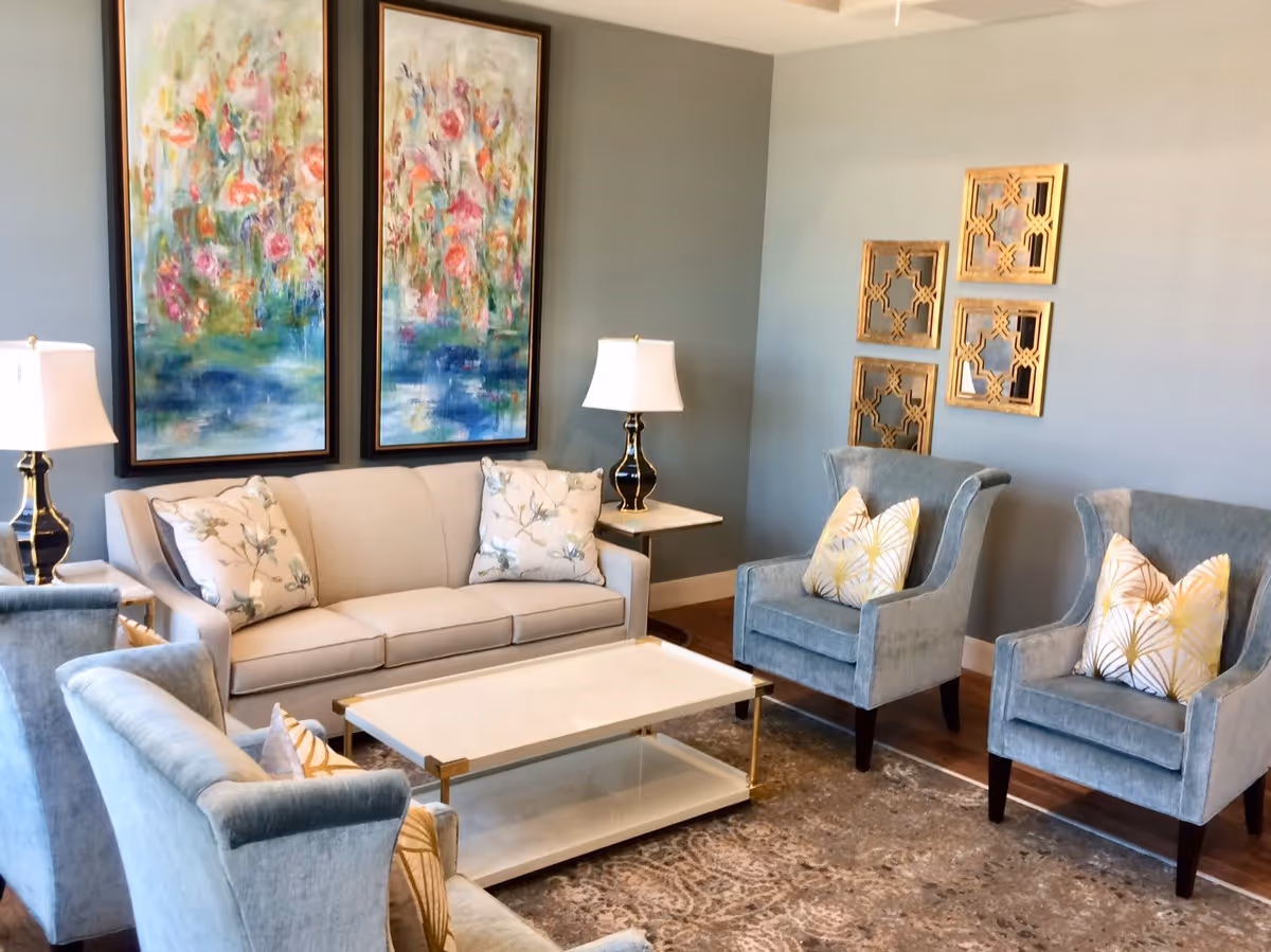 A cozy living room with a beige sofa adorned with floral pillows, two blue armchairs with patterned cushions, a white coffee table with gold accents, two table lamps with white shades, and colorful floral paintings on the wall.