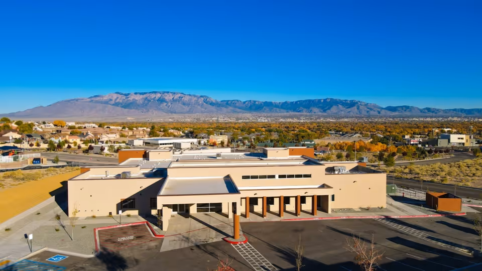Aerial view of a single-story beige assisted living building with a parking lot in front and mountains in the background.