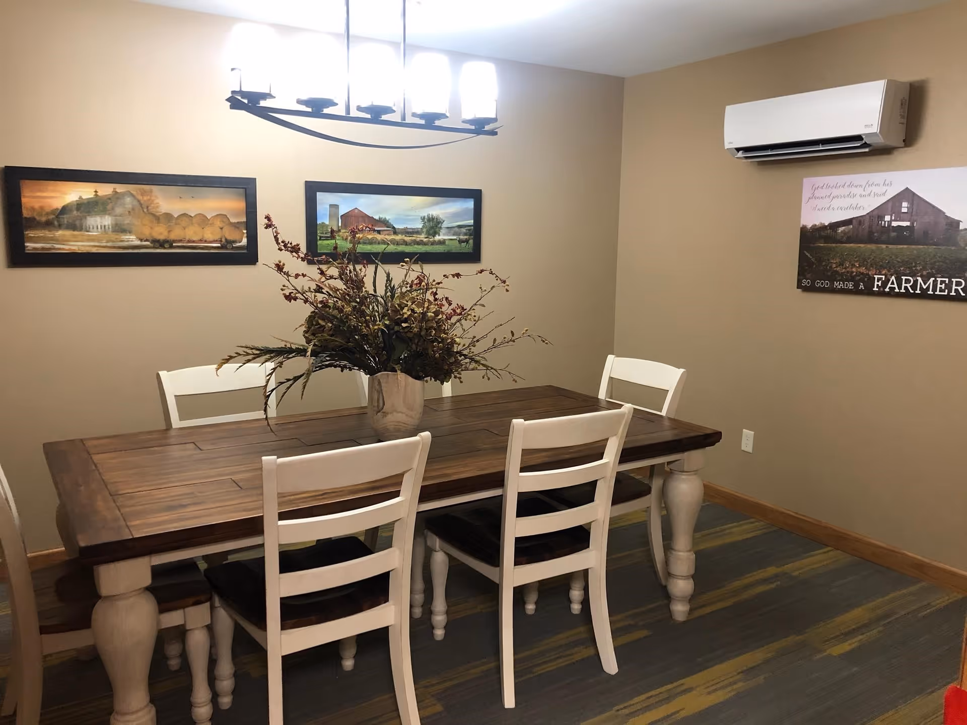 A dining room with a wooden table and four white chairs. A vase with a floral arrangement sits in the center of the table. Three framed pictures hang on the beige walls, and a modern light fixture is mounted on the ceiling above the table. An air conditioning unit is installed on the wall.