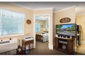 Interior view of a living area in Meadow Creek Village Assisted Living showing a window with blinds and a small table with a typewriter and other items. A doorway leads to a bedroom with a bed and framed pictures on the wall. A TV on a wooden stand displays an outdoor scene, and a decorative fish wall hanging is above the TV.