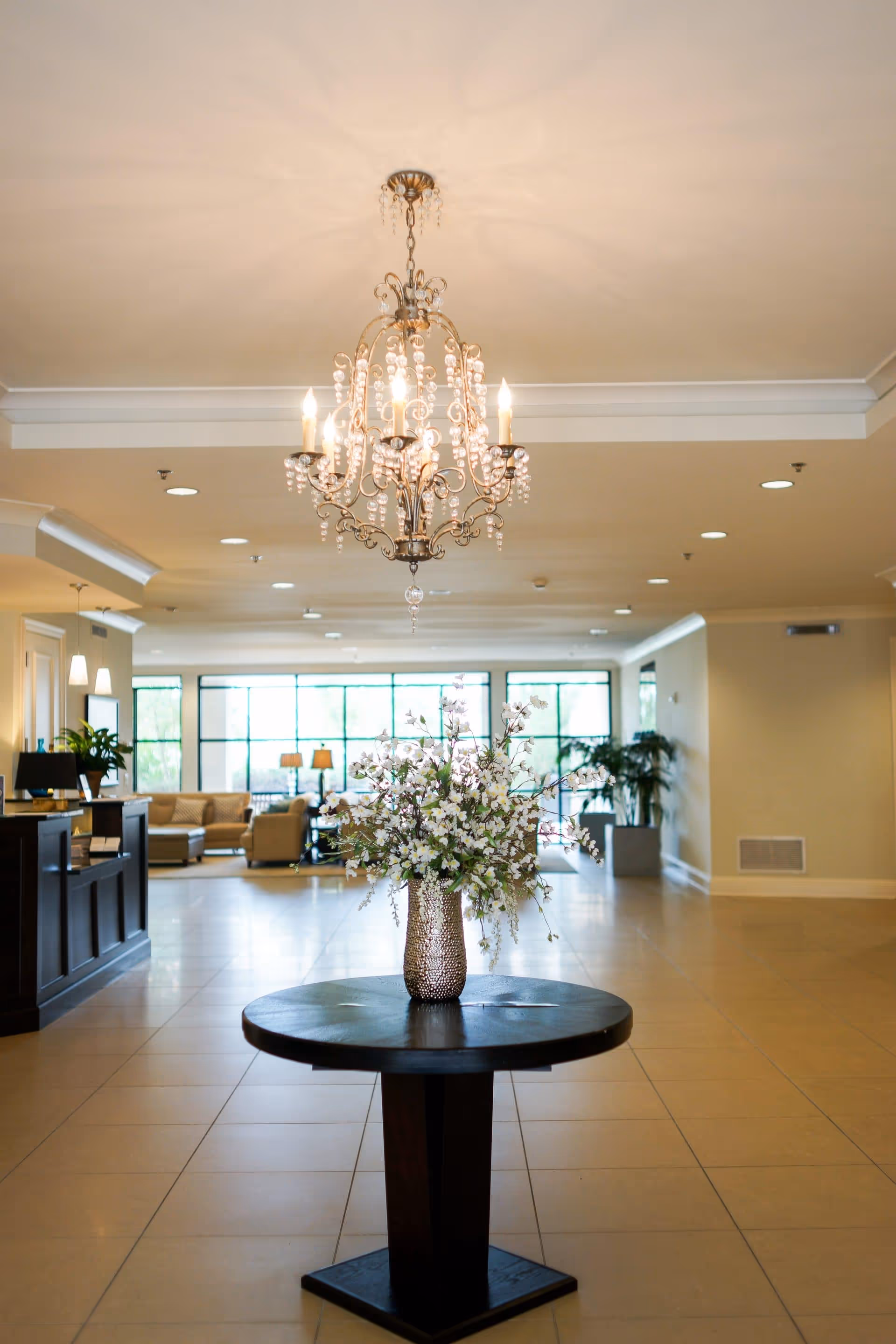 A spacious and well-lit lobby area with a round wooden table in the center holding a vase filled with white flowers. Above the table hangs an elegant chandelier with multiple lit bulbs. In the background, there is a seating area with beige sofas and large windows letting in natural light.