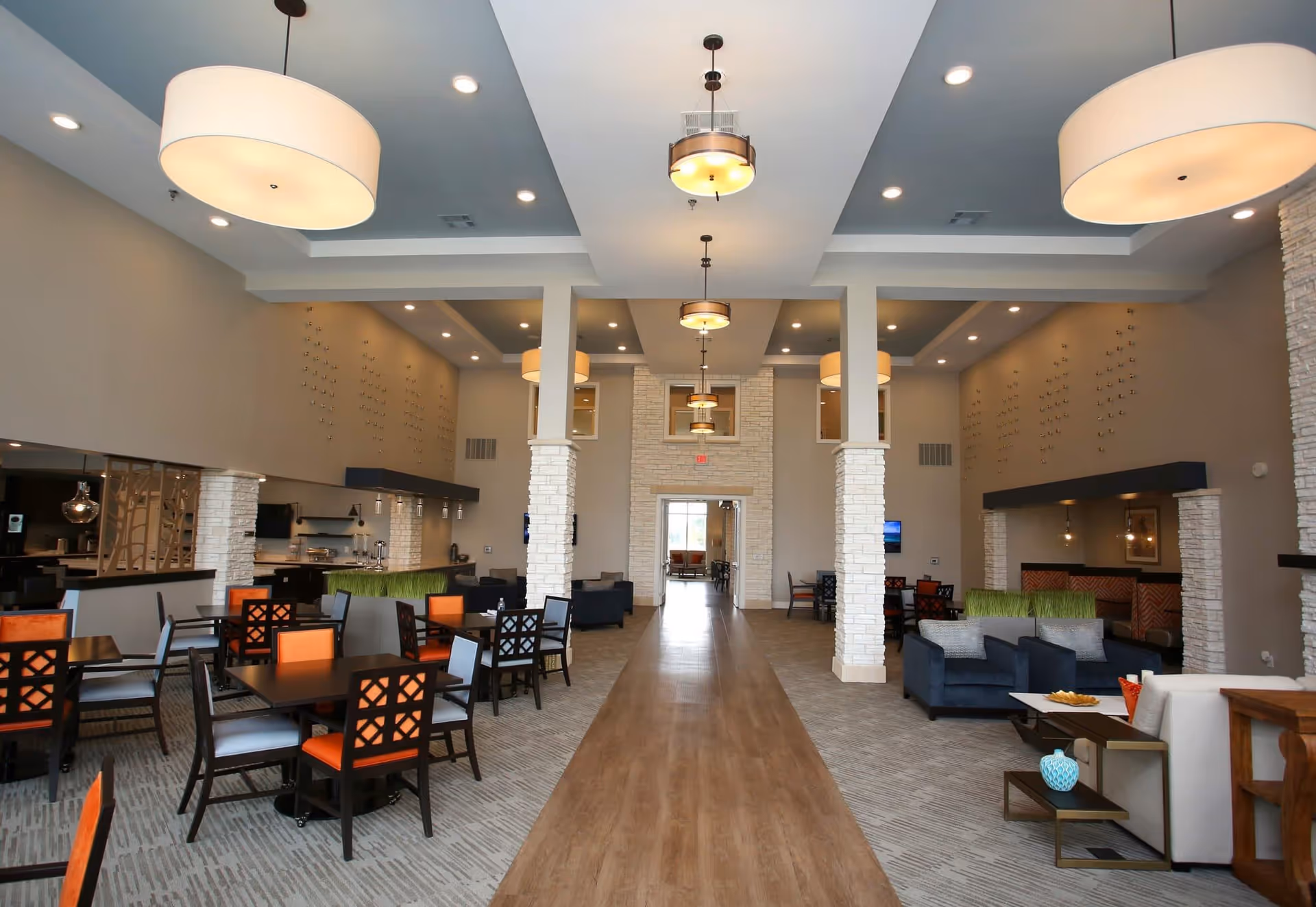 Spacious and modern common area in a senior living facility featuring a dining section with tables and chairs on the left, a lounge area with sofas and armchairs on the right, large pendant lights hanging from the ceiling, and a wide wooden walkway down the center leading to a doorway at the back.