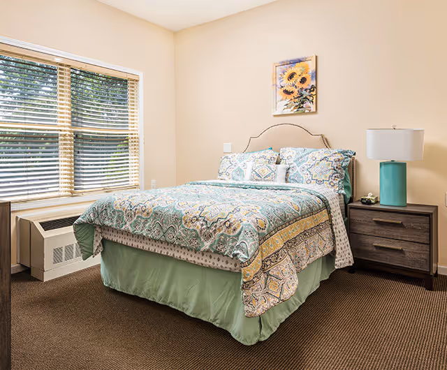 Bright bedroom with a bed dressed in patterned teal and yellow bedding, a nightstand with a lamp, a large window with blinds, and floral wall art.