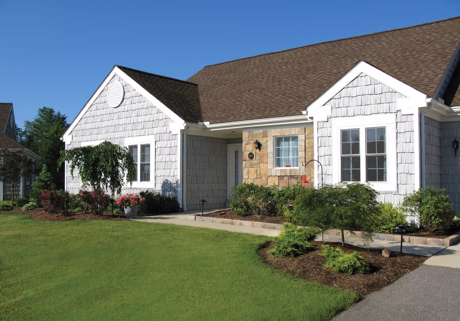 Exterior view of a single-story residential building with gray shingle siding, white trim, and a brown shingled roof. The front entrance is recessed with a stone facade around the door and window. There is a well-maintained lawn and landscaped garden beds with shrubs and flowering plants under a clear blue sky.