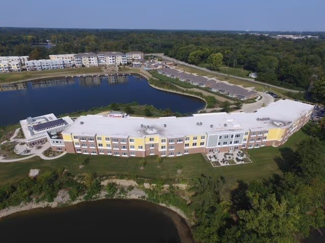 Aerial view of Traditions at Solana senior living facility showing a large, curved multi-story building with a white roof, situated next to a pond and surrounded by greenery and trees.