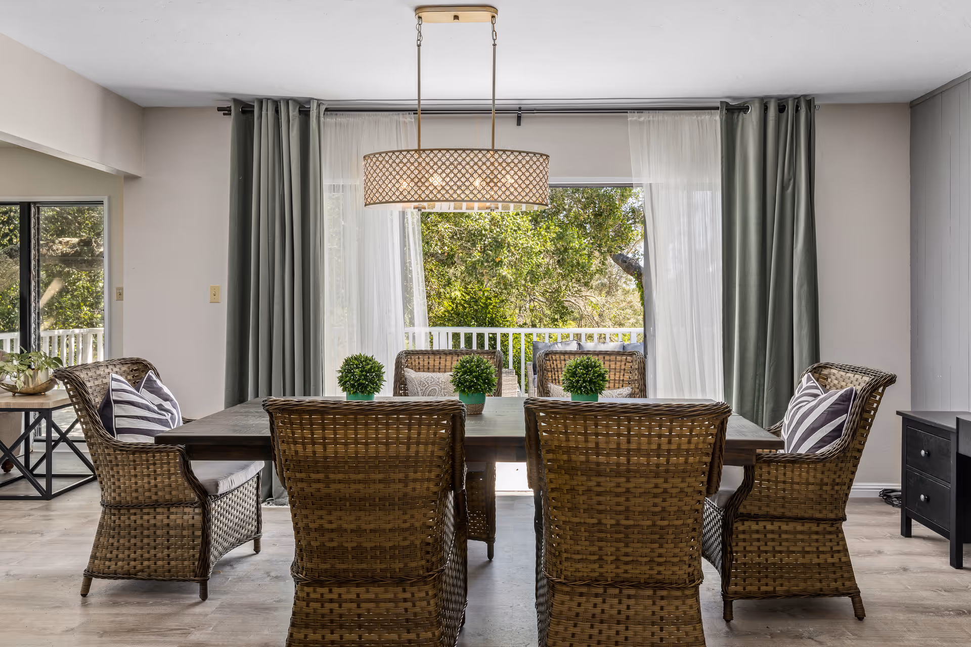 A dining room with a rectangular wooden table surrounded by six wicker chairs with cushions. Three small potted plants are placed on the table. Behind the table, large windows with sheer white curtains and green drapes reveal a view of trees and a white railing outside. A decorative pendant light hangs above the table.