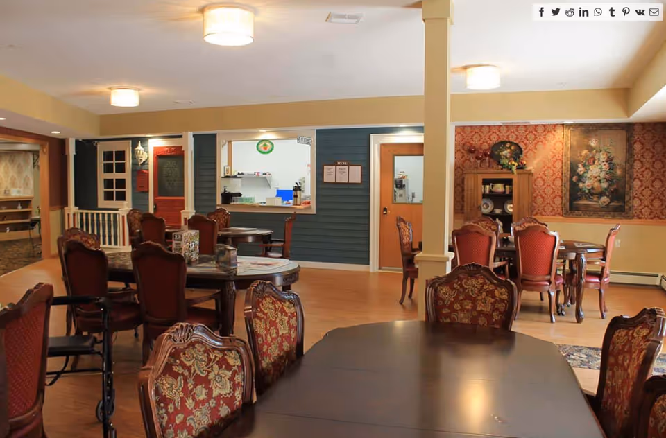 A dining room in a senior living facility with multiple wooden tables and ornate red upholstered chairs. The room has warm lighting, wooden flooring, and decorative wallpaper with floral paintings. There is a serving window and a door on the far wall, with a menu displayed beside the window.
