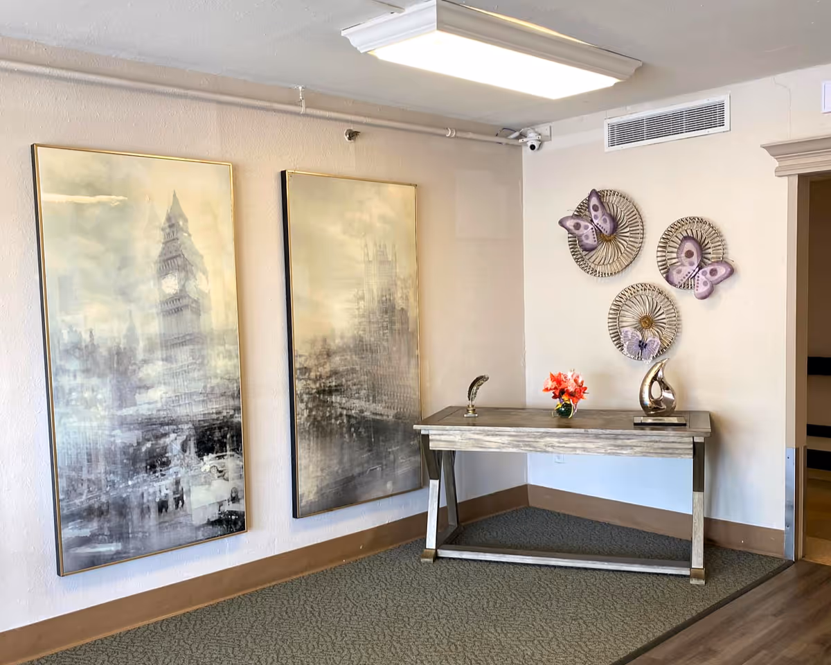 An interior corner of a room with two large framed abstract paintings on the left wall depicting cityscapes. A wooden console table is positioned against the adjacent wall, decorated with a small vase of red flowers, a decorative sculpture, and a feather-shaped ornament. Above the table, three circular wall decorations with purple butterflies are mounted. The floor is carpeted and the walls are painted light beige.
