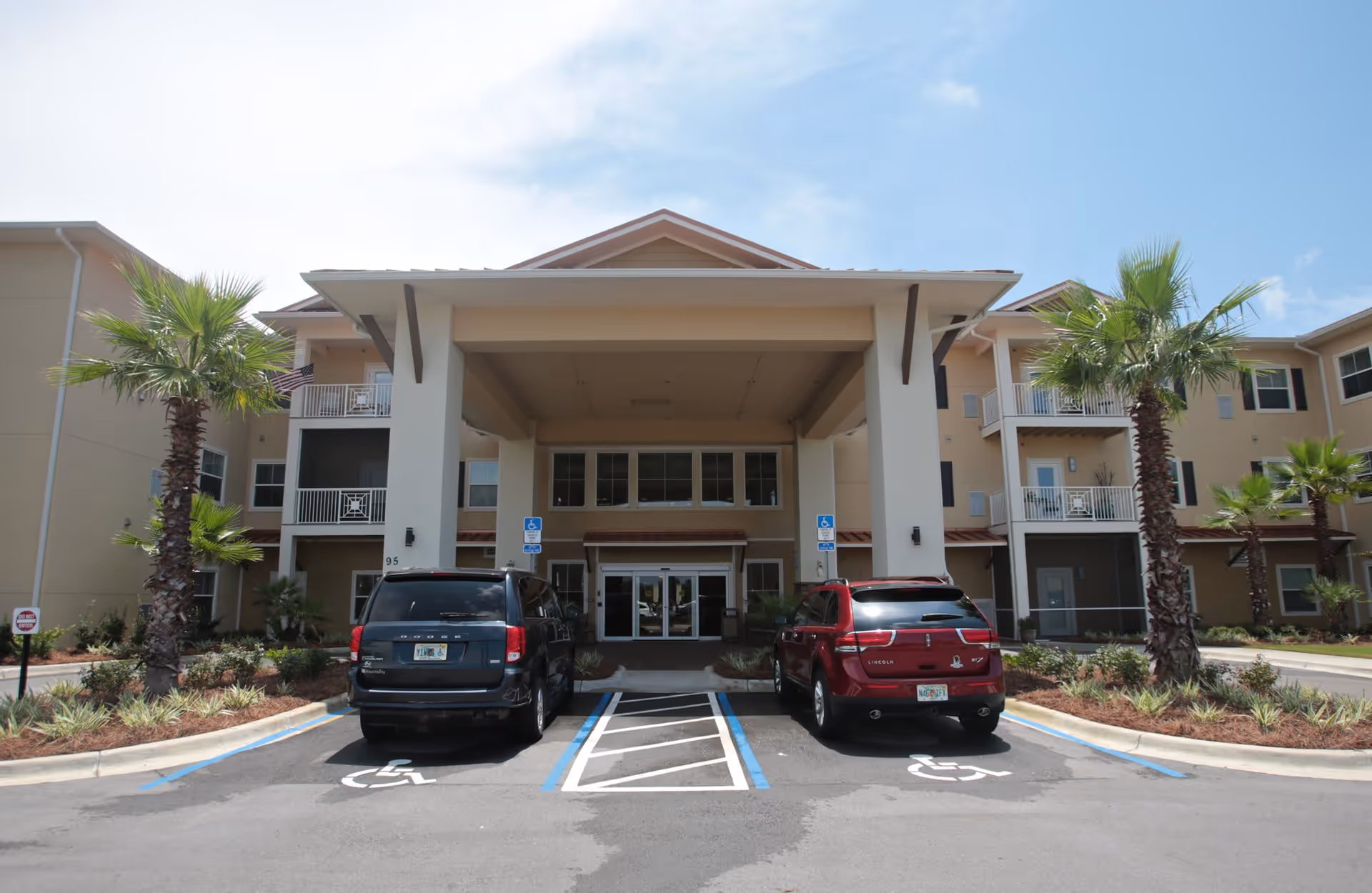 Front exterior view of a senior living facility named Superior Residences of Panama City Beach, showing the entrance with a covered drop-off area, two parked cars in handicapped parking spaces, palm trees, and a clear blue sky.