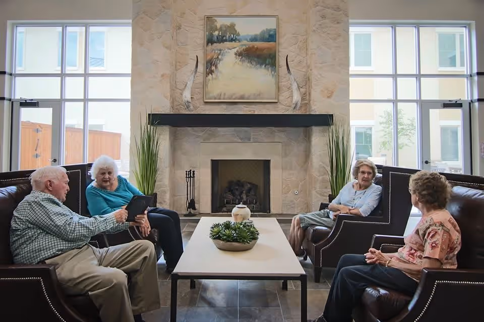 Four elderly people sitting on leather chairs around a rectangular coffee table in a well-lit living room with large windows and a stone fireplace. The room is decorated with plants and a landscape painting above the fireplace.