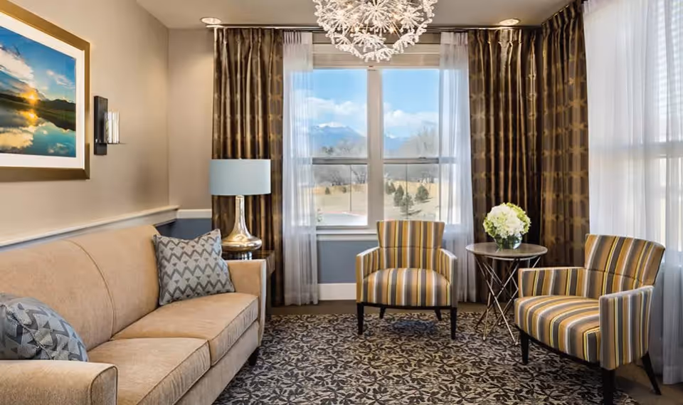 A cozy living room with a beige sofa adorned with patterned pillows, two striped armchairs, a small round table with a flower arrangement, a lamp on a side table, and a large window with sheer and patterned curtains letting in natural light.