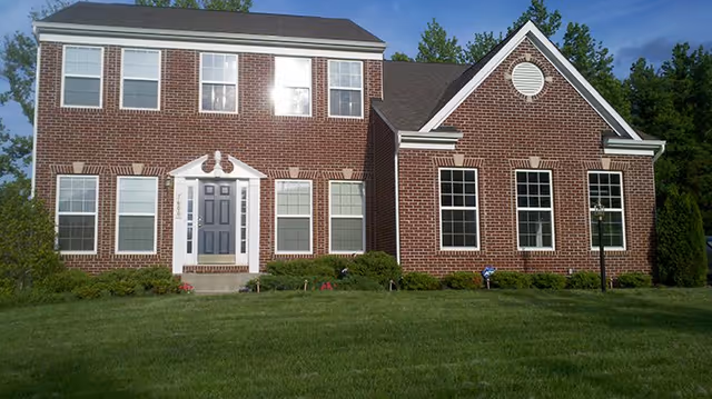 Front exterior view of a two-story brick building with multiple windows, a gray front door with white trim, and a well-maintained green lawn in front. The building is surrounded by trees and shrubs under a clear blue sky.