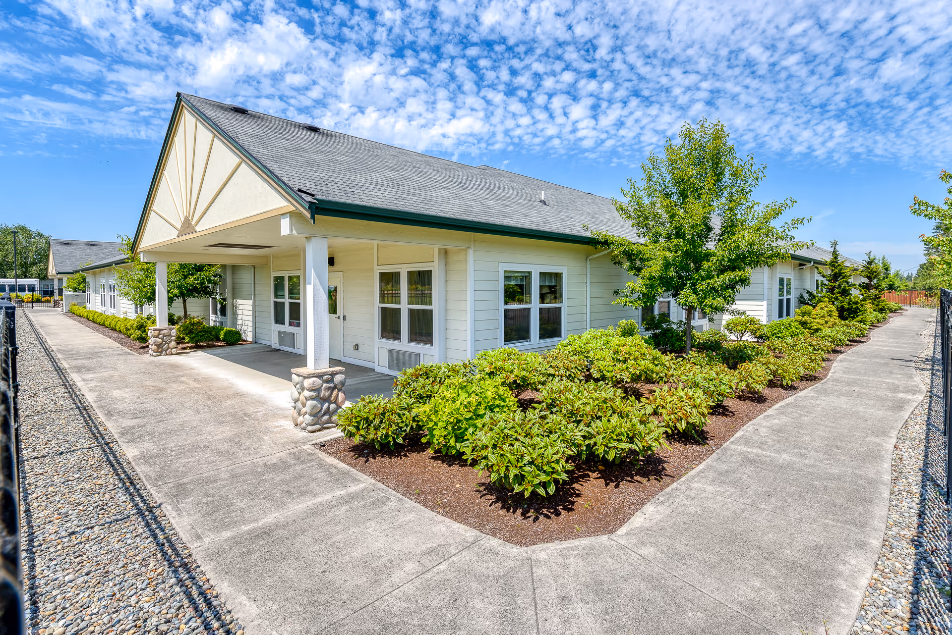 Exterior view of a single-story assisted living facility building with a covered entrance, surrounded by well-maintained landscaping including bushes and trees, under a partly cloudy blue sky.
