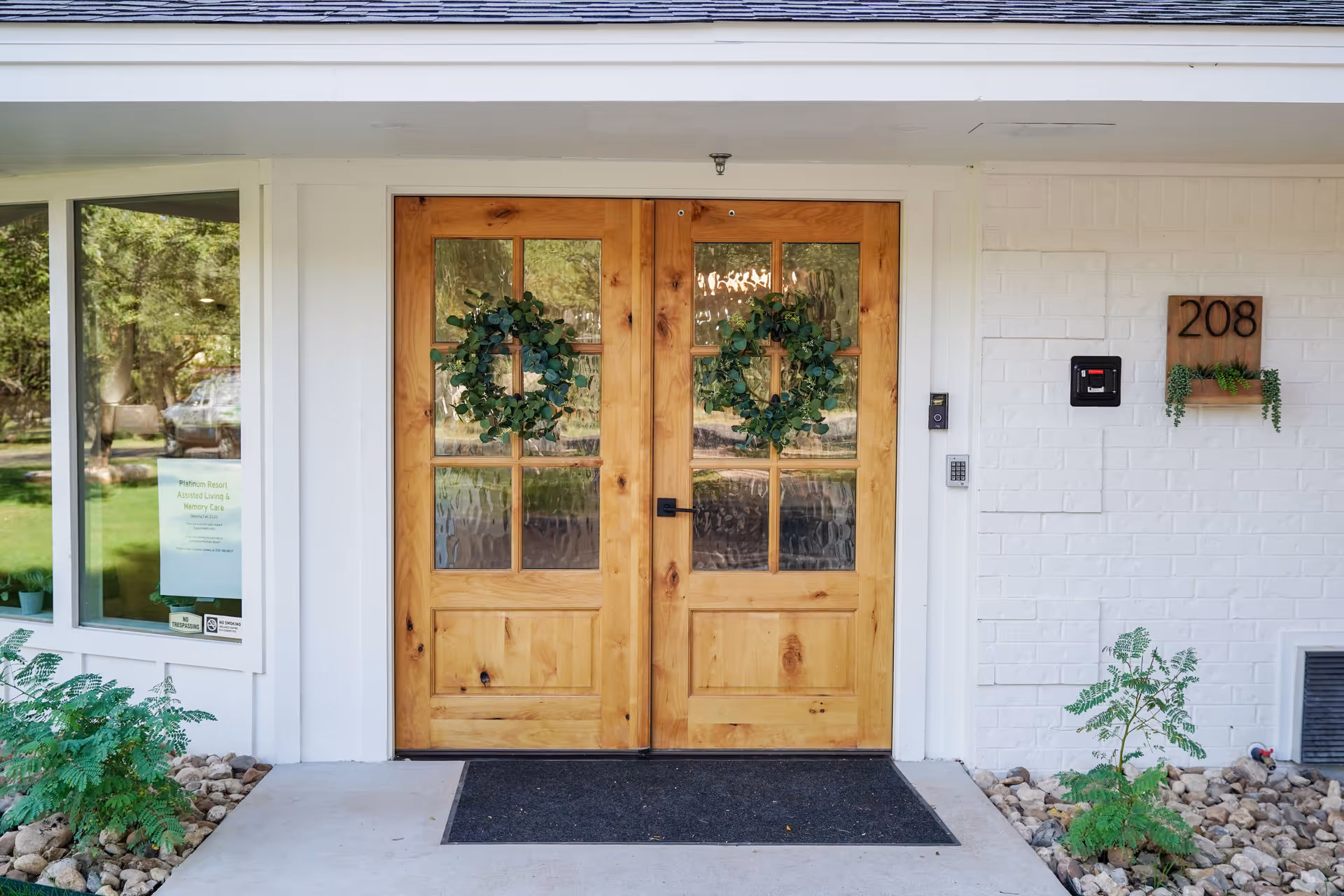 Front entrance of a building with double wooden doors decorated with green wreaths. The building has white brick walls, a window on the left side with a sign visible inside, and a wooden plaque with the number 208 and small plants on the right side. There are small plants and rocks along the base of the wall.