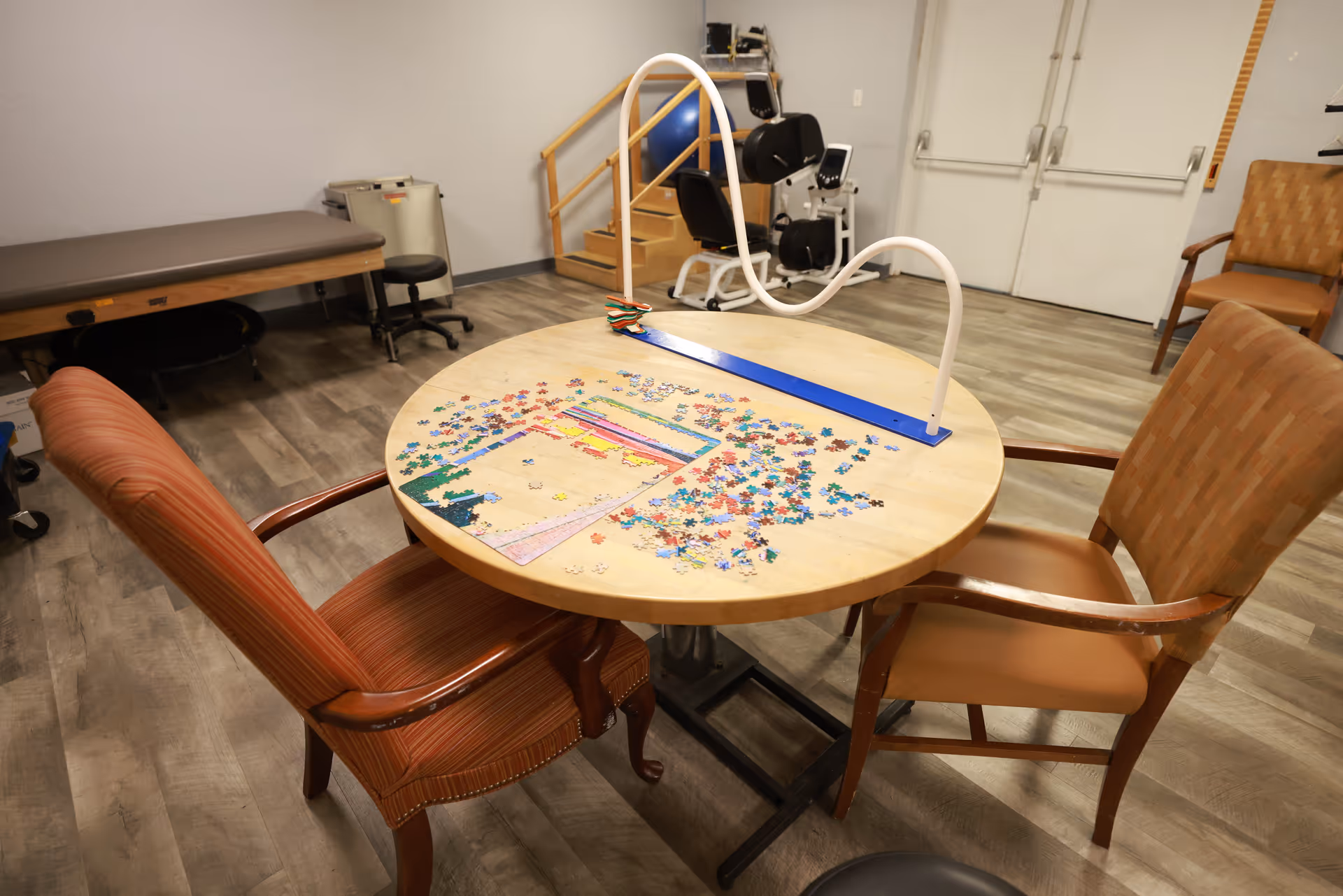 A therapy or activity room with a round table in the center holding a partially completed jigsaw puzzle and a puzzle piece holder. Two chairs are positioned around the table. In the background, there is exercise equipment, a padded bench, and a set of stairs with a handrail. The floor is covered with wood-patterned vinyl flooring and the walls are plain light-colored.