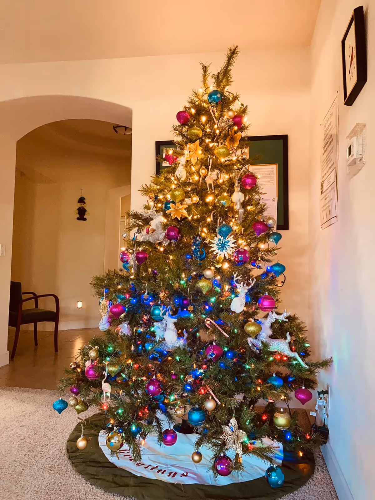 A decorated Christmas tree inside a room with colorful ornaments including pink, blue, gold, and green baubles, white reindeer figures, and multicolored string lights. The tree skirt is green and white with red text. In the background, there is a chair, an archway leading to another room, framed pictures on the wall, and a thermostat.