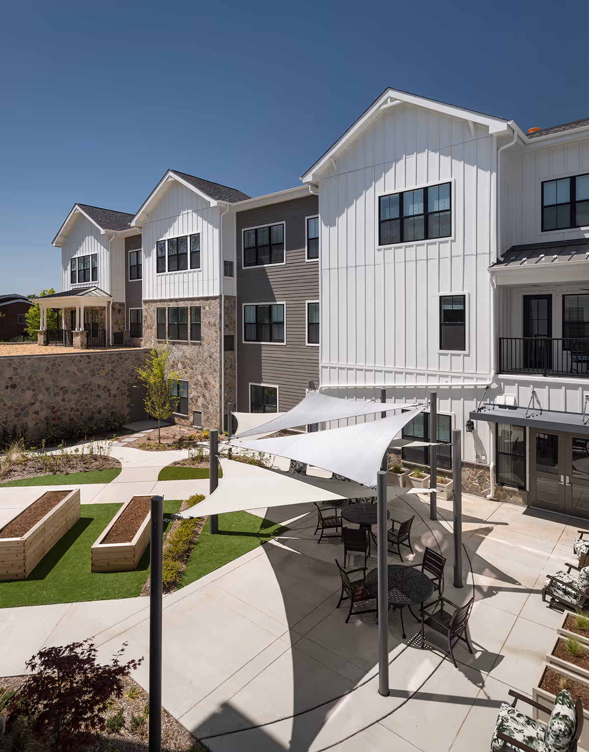 Outdoor courtyard area of a senior living facility with white shade sails over round tables and chairs, raised garden beds, and a multi-story building with white and gray siding and stone accents under a clear blue sky.