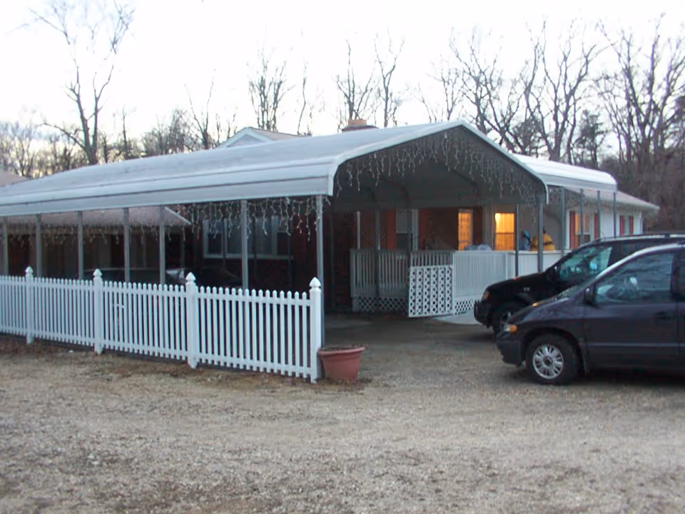 Front view of a single-story home with a covered carport, white picket fence, and two parked cars.