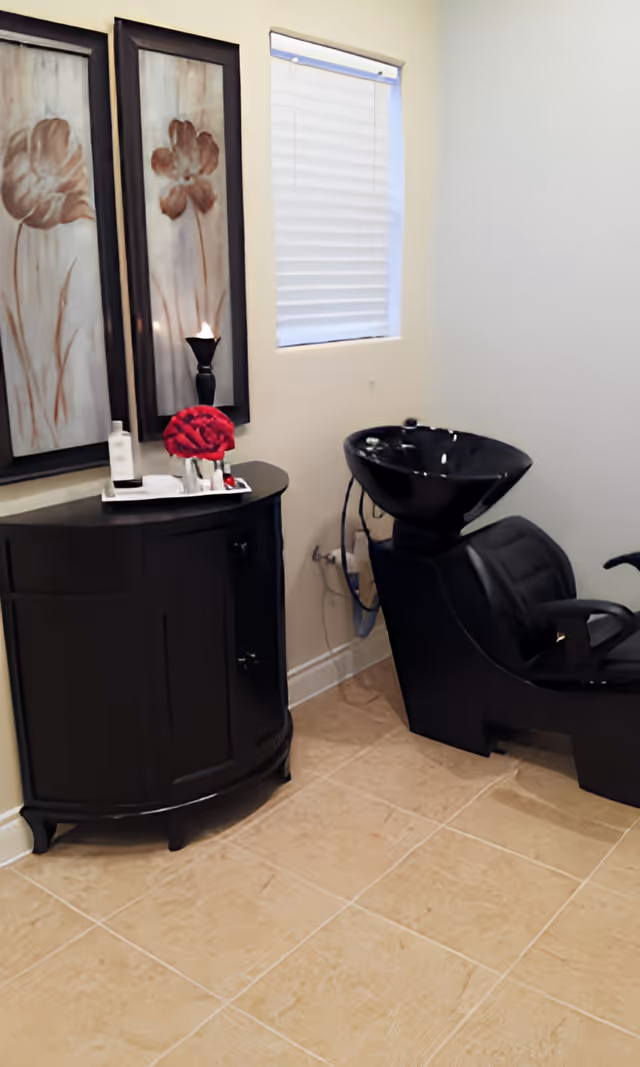 A small room with a black salon shampoo chair and sink, a black curved cabinet with a tray holding a bottle and red flowers, two framed floral artworks on the wall, and a window with closed blinds.