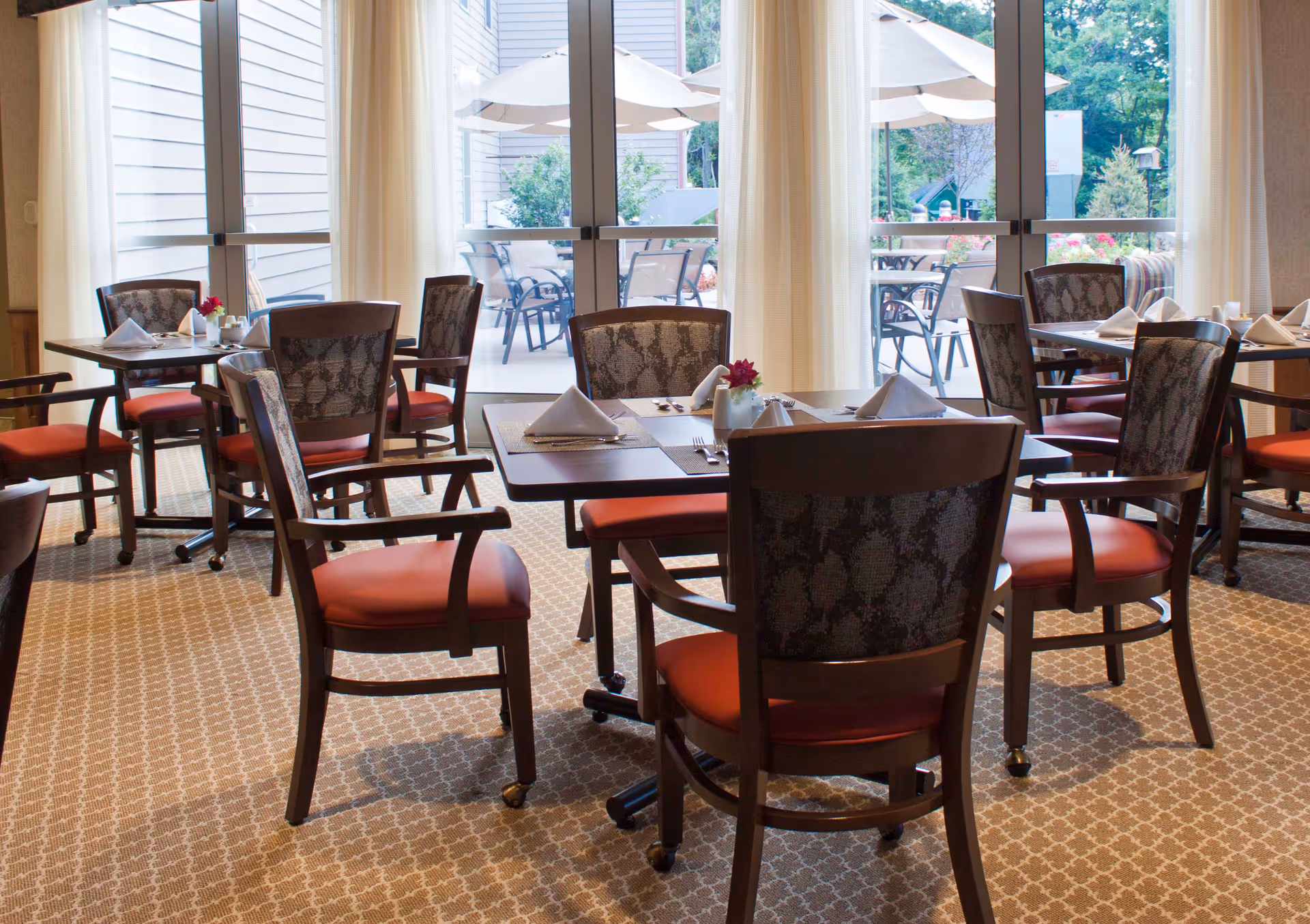 Dining area with several tables and chairs arranged neatly, each table set with napkins, cutlery, and small flower vases. Large glass doors in the background open to an outdoor patio with umbrellas and additional seating, surrounded by greenery.